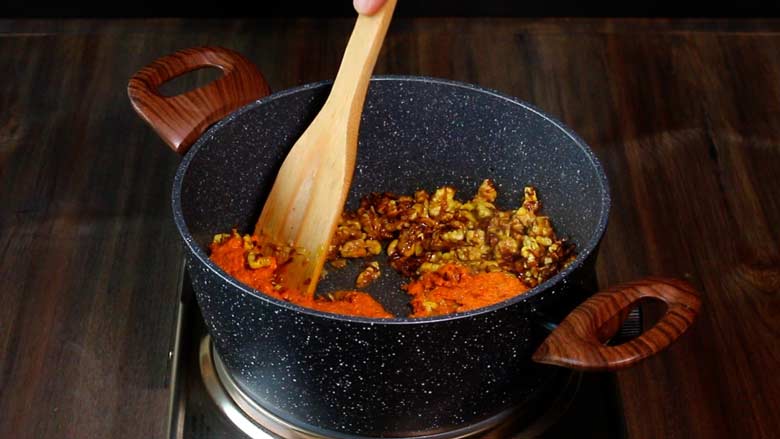 A wooden spatula is seen stirring wet masala paste into the walnut mixture in a grey and white speckled non stick pan on an electric stove on a wooden counter - Malvani Green Peas and Walnut Curry Recipe